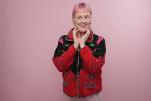 A white woman with a short pink mullet is looking to her right, she has a cheeky grin and her hands are slightly touching her face. She is wearing a red and black knitted embroidered jacket.